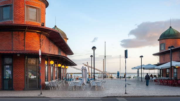 Un edificio frente al agua con fachada de ladrillo, varias áreas de comedor con mesas al aire libre y una estatua en el centro.
