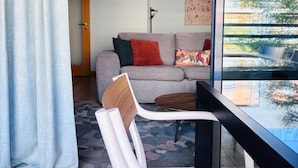 Un salón moderno con un sofá, una silla, una mesa y un amplio ventanal con vistas a edificios y árboles.