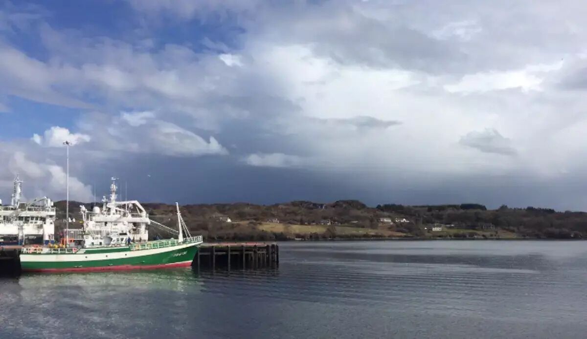 En grön och vit båt låg förtöjd vid en brygga, och vattnet var stilla. I bakgrunden fanns ett kuperat landskap.