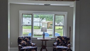 A room with two floral armchairs, a small table with a lamp, and a view of a house and yard through large windows. - Restful Farm Retreat (Cincinnati)