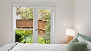 Une chambre à coucher comprenant un lit, une table de chevet, une lampe et une vue sur une clôture en bois et de la verdure, visible à travers une porte-fenêtre coulissante.