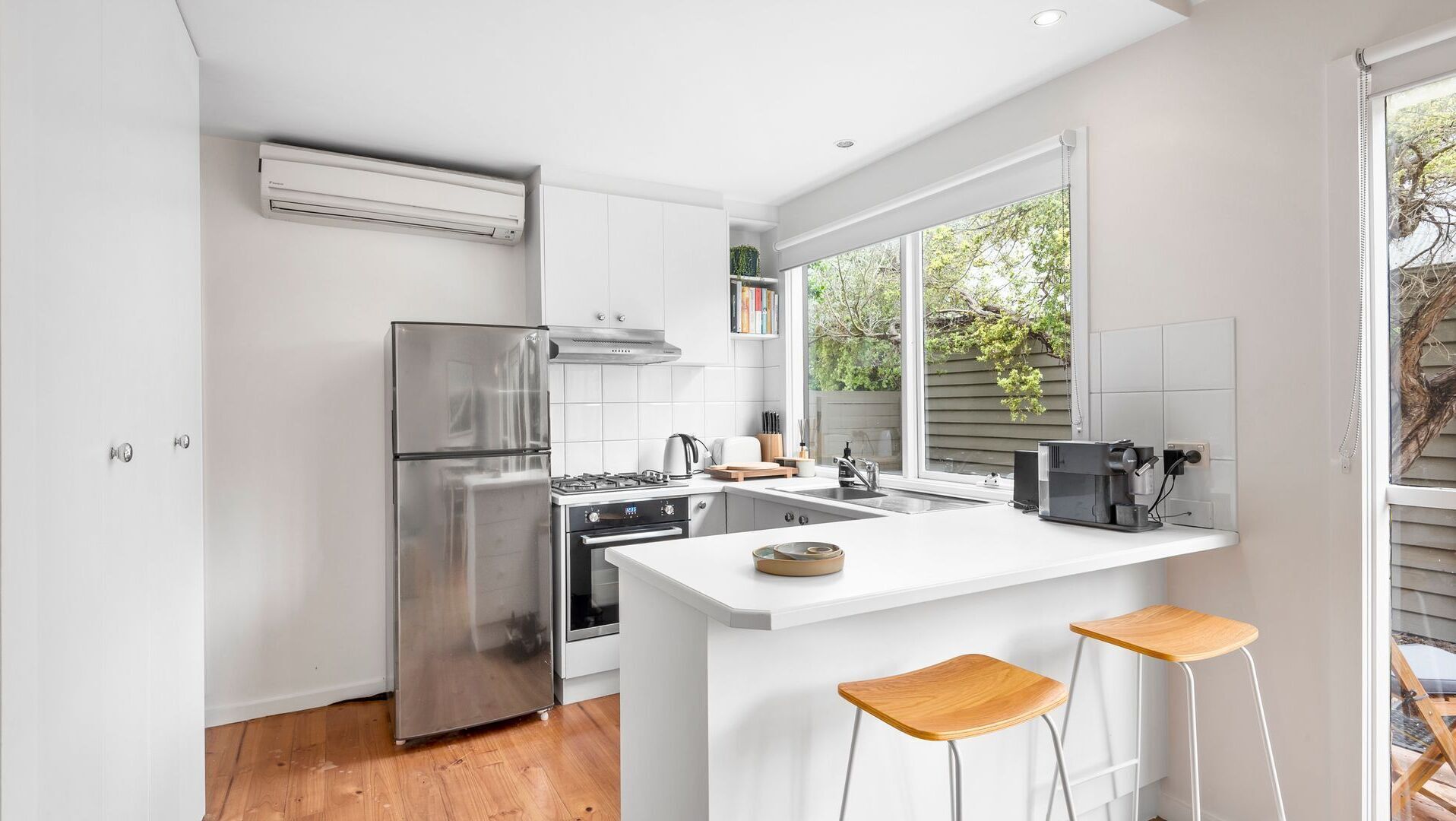 A modern kitchen with white cabinetry, stainless steel appliances, a large window, and a small island with bar stools.