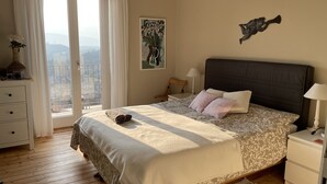 Et soveværelse med en stor seng, et natbord, en kommode, et vindue med gardiner og et billede på væggen.