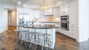 A modern kitchen with white cabinetry, stainless steel appliances, and a central island with bar stools. - Bella Luna 504 by Liquid Life (Orange Beach)