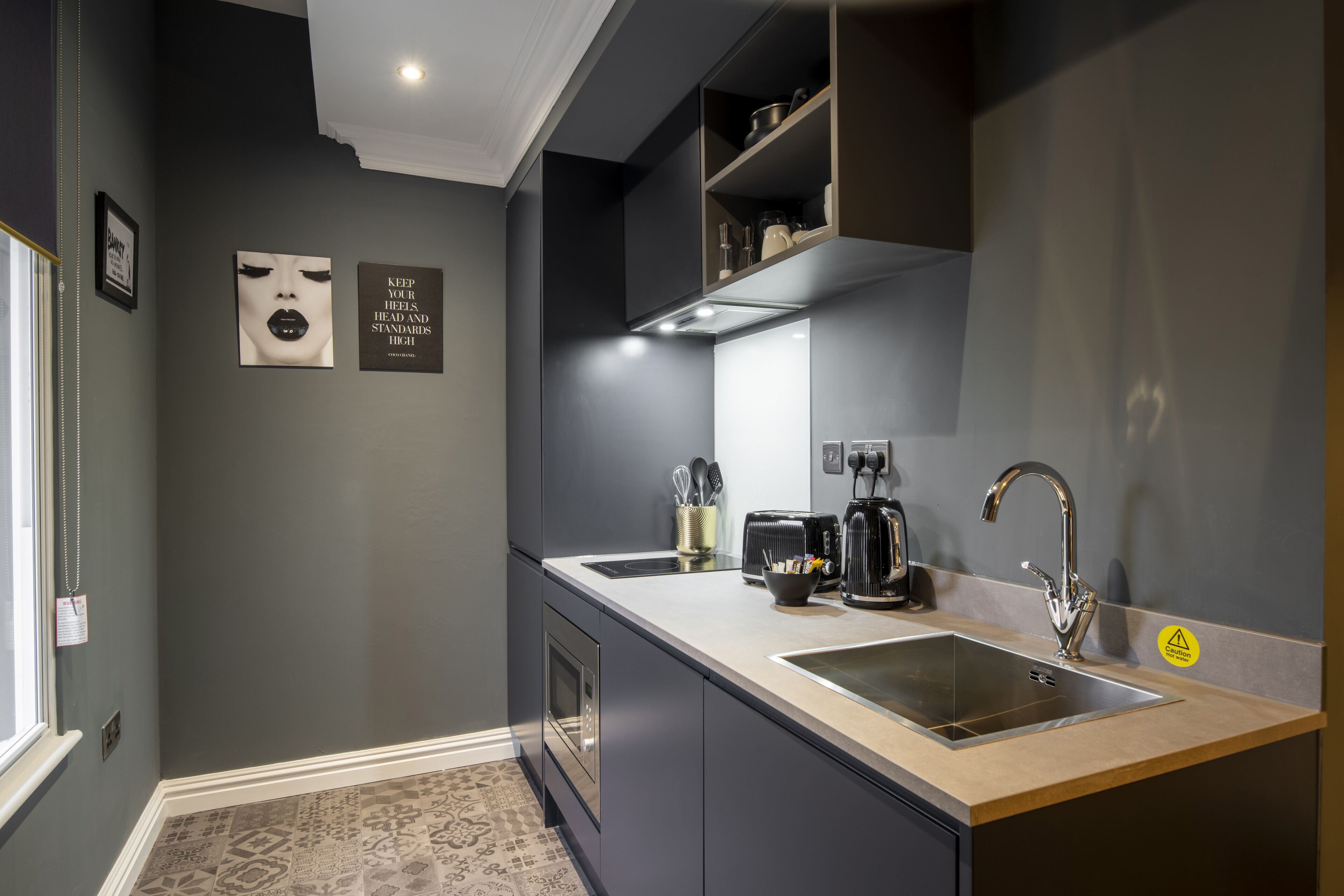 A modern kitchen with dark cabinetry, a stainless steel sink, and a built-in range hood. There are two framed pictures on the wall and a small shelf with kitchen items.