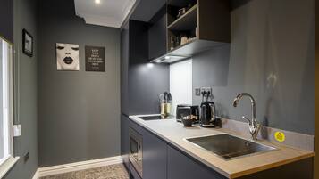 A modern kitchen with dark cabinetry, a stainless steel sink, and a built-in range hood. There are two framed pictures on the wall and a small shelf with kitchen items.