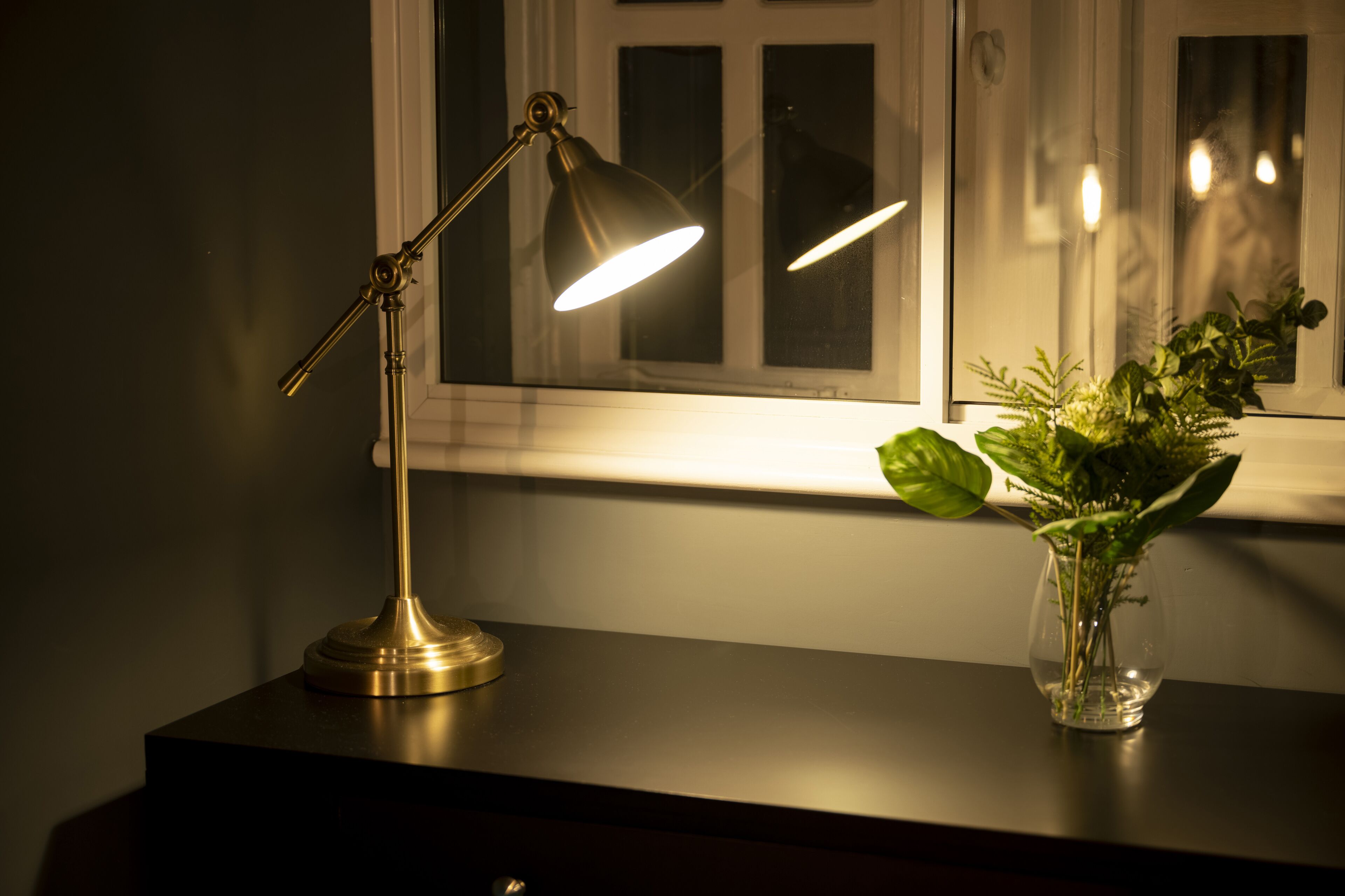 En bordslampa och en vas med växter står på ett bord nära ett fönster.