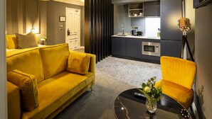 Ein modernes Wohnzimmer mit einem gelben Sofa, einem runden Couchtisch und einem gelben Sessel. Im Hintergrund ist eine Küchenzeile mit Kühlschrank, Herd und Spüle zu sehen.