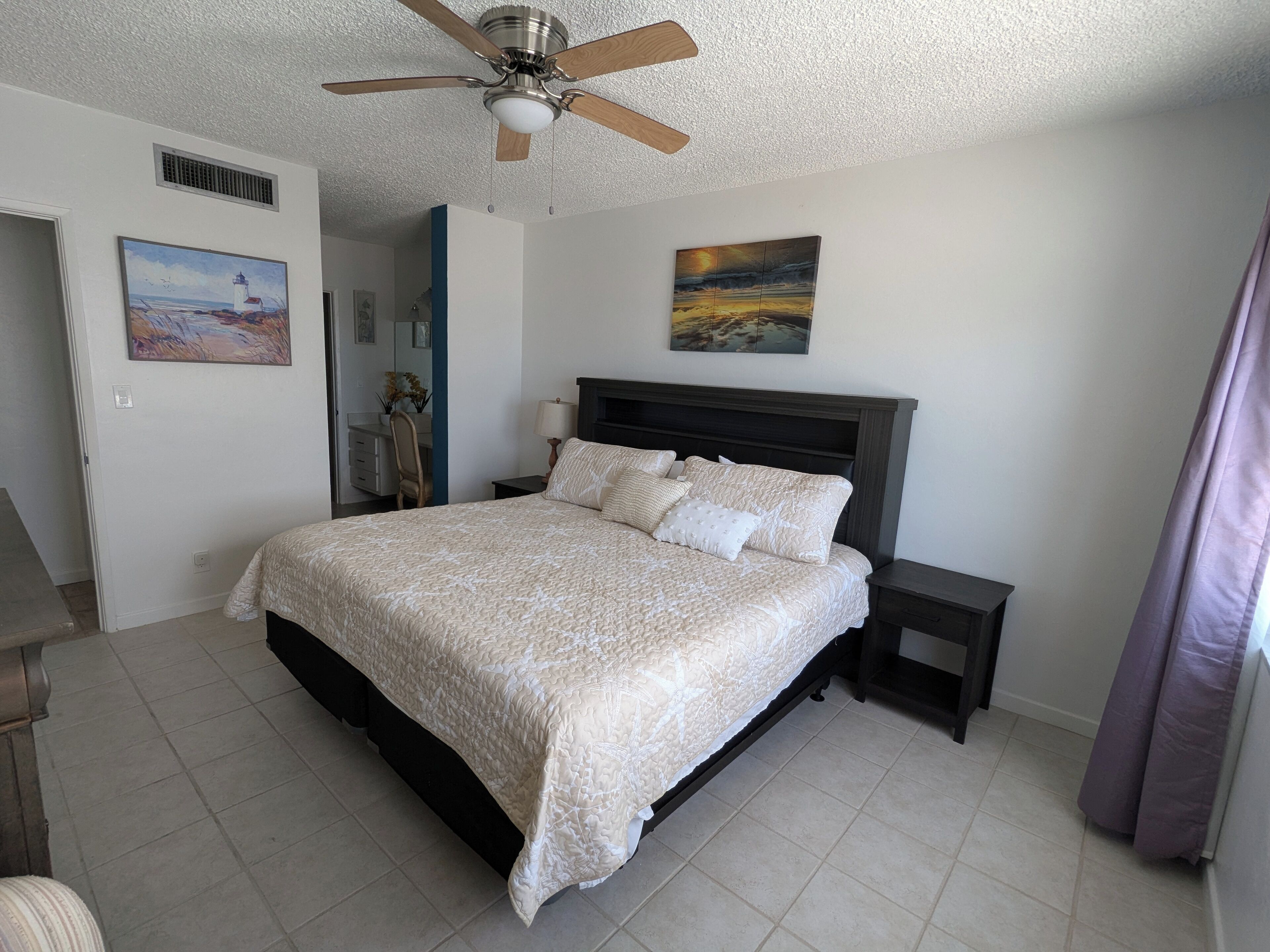 A bedroom with a bed, a ceiling fan, a nightstand, a lamp, a painting, and a door.