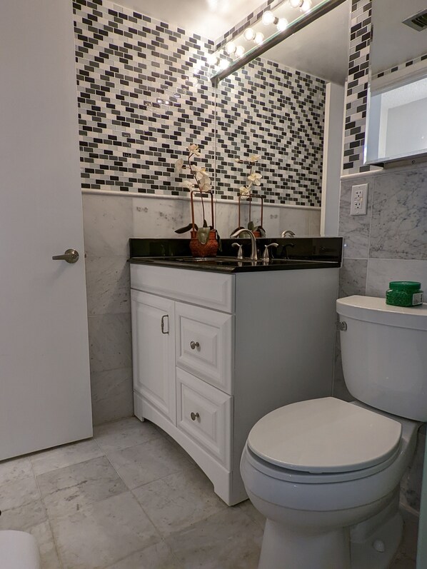 Een badkamer met een betegelde spatwand, een wit wastafelmeubel met lades, een toilet en een spiegel met verlichting erboven.