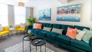 A modern living room with a teal sofa, yellow chairs, and a patterned rug. There are two paintings on the wall, one depicting a cityscape and the other beach houses. - Station Lodge By My Getaways (Brighton)