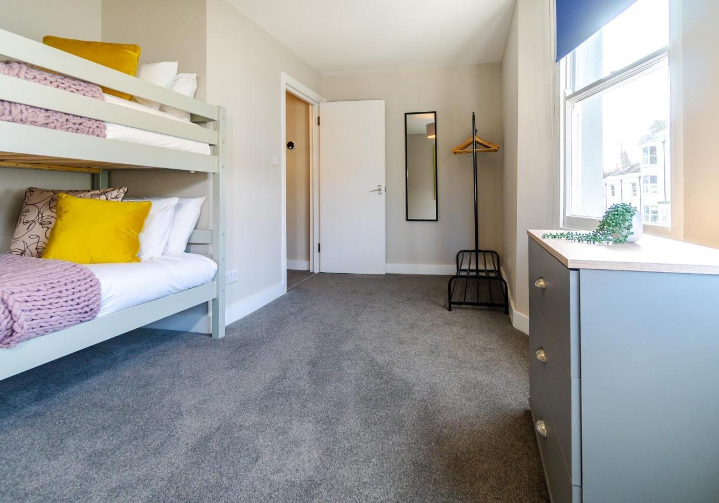 Une chambre avec un lit superposé, un bureau, un miroir et une fenêtre donnant sur des immeubles.