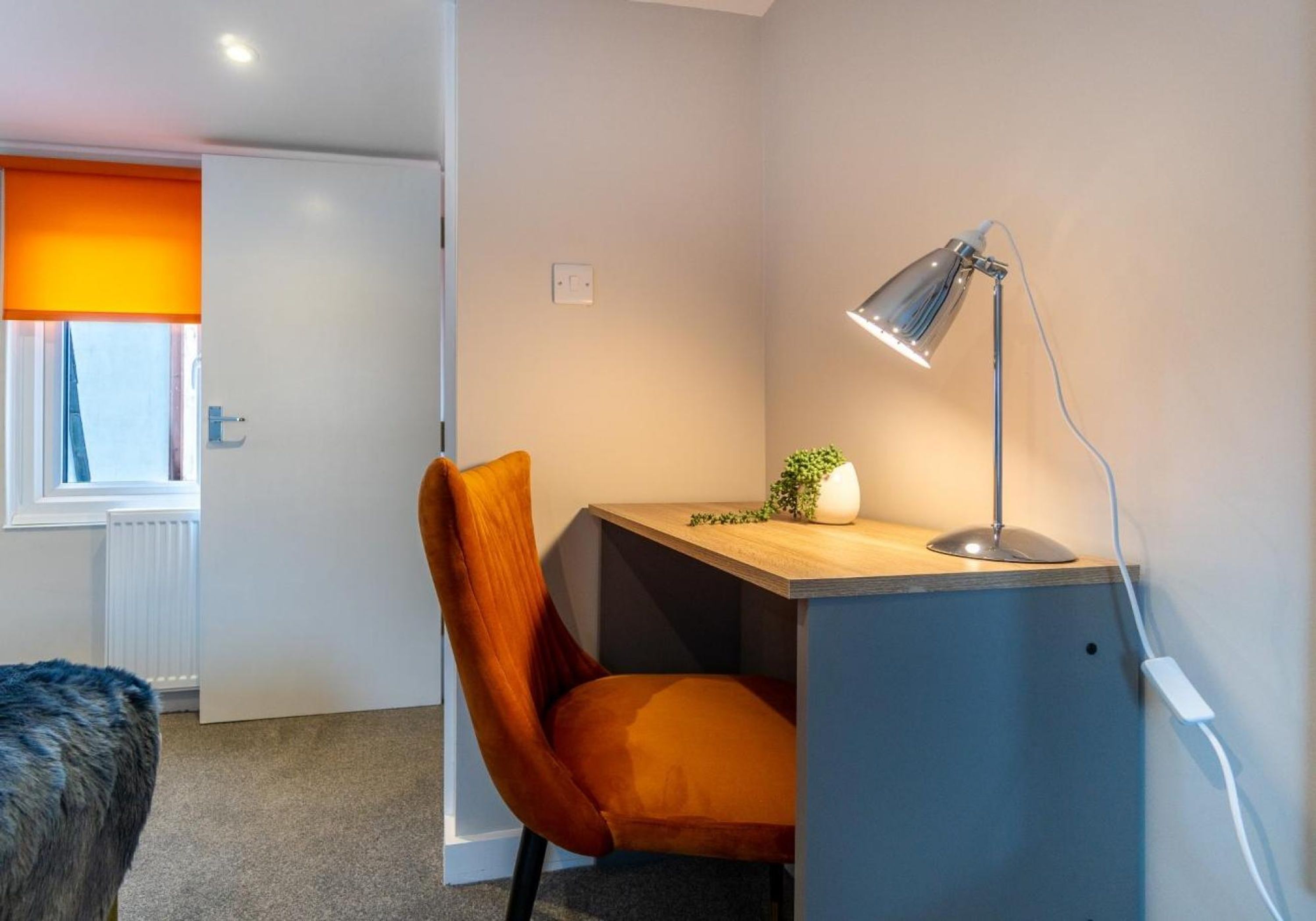 Een kleine, goed verlichte kamer met een bureau, stoel en lamp. Een radiator is zichtbaar op de achtergrond, en een deur staat open naar buiten.