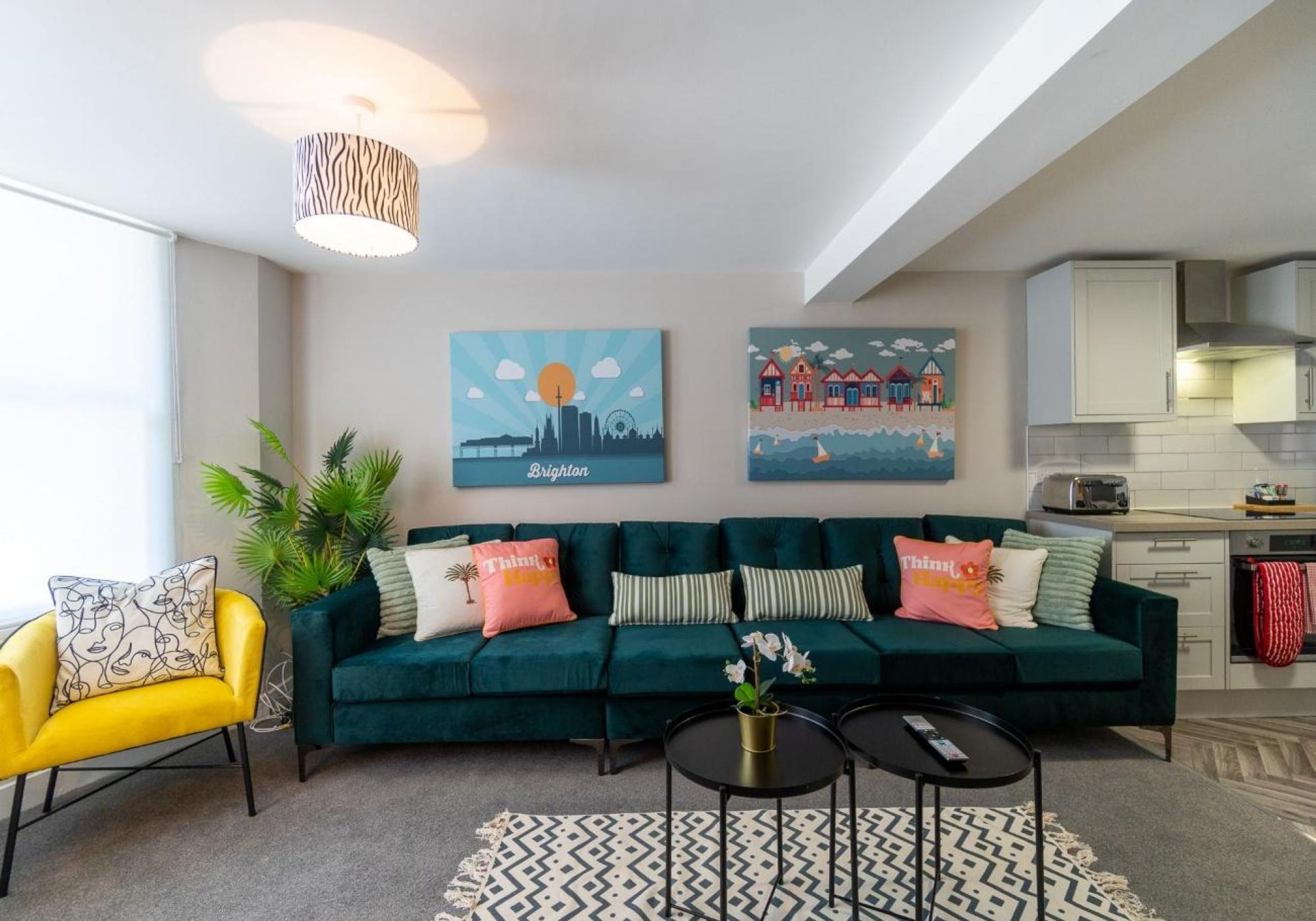 Un salon moderne avec un canapé vert, un fauteuil jaune et un tapis à motifs. Deux tableaux ornent le mur : l’un représente un paysage urbain et l’autre, une scène côtière.