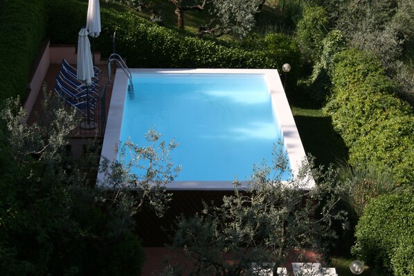 A rectangular swimming pool surrounded by lush greenery, with a patio area featuring a chair and umbrella. - Independent Portion of Luxury Villa with Pool close to Assisi (Umbria)