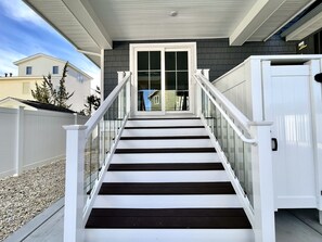 Un escalier mène à une maison à l’extérieur blanc et aux bardages sombres, dotée d’une porte d’entrée en verre et entourée d’une clôture blanche.