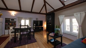 A cozy living room with a dining area, wooden beams on the ceiling, a TV, and a window with curtains.