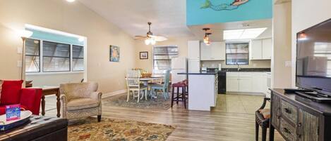 A spacious living room with a large rug, a sofa, a dining area with chairs, a kitchen with white cabinets, and a ceiling fan.