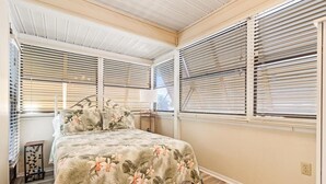 A bedroom with a bed, two windows with blinds, a small bedside table, and a nightstand.
