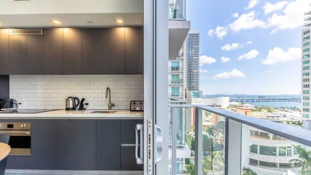A modern kitchen with dark cabinets, white tiles, and a view of a cityscape through a balcony. - Rooftop Pool & Ocean Views Near Bayside (Miami)