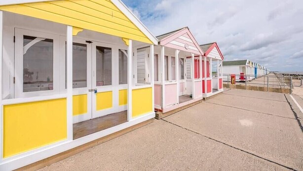 A row of beach huts with white and yellow exteriors, each with a small porch and windows. - Little Hotsie, Southwold (Southwold)