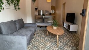 Una sala de estar moderna con un sofá gris, una mesa de centro redonda de madera, baldosas de suelo con diseño, una lámpara colgante y un televisor sobre un mueble blanco.