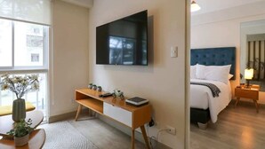 A modern hotel room with a large flat-screen TV mounted on the wall, a wooden desk with a laptop, a bedside table with a lamp, and a bed with a blue headboard. - w* | 1BR near Central Restaurant in Barranco (Barranco)