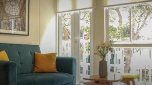 A living room with a teal sofa, a yellow pillow, a framed picture on the wall, and a window with a view of trees. - w* | 1BR near Central Restaurant in Barranco (Barranco)