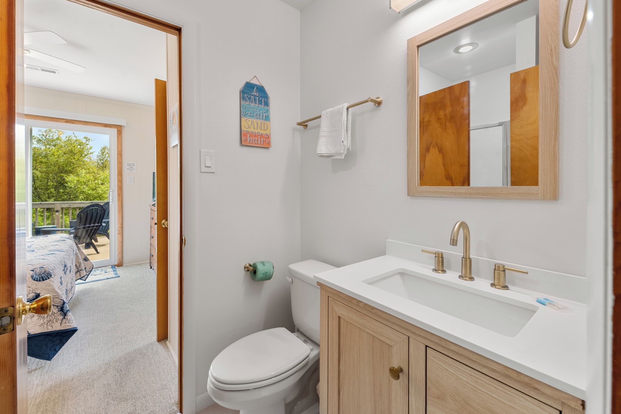 A bathroom with a white sink, gold faucets, and a wooden cabinet. There is a toilet, a towel rack, and a sign on the wall.