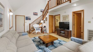 Een ruime woonkamer met een hoge plafond, een houten trap, een zithoek, een salontafel en een televisie.