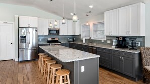 A modern kitchen with a central island, bar stools, stainless steel appliances, and a breakfast bar. - Salty Sunsets: Hot Tub | 2 Kitchens | Pool Access (Corolla)