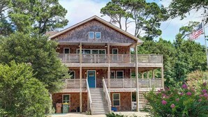 A two-story wooden cabin with a balcony on the upper level, surrounded by trees and a flag. - Salty Sunsets: Hot Tub | 2 Kitchens | Pool Access (Corolla)