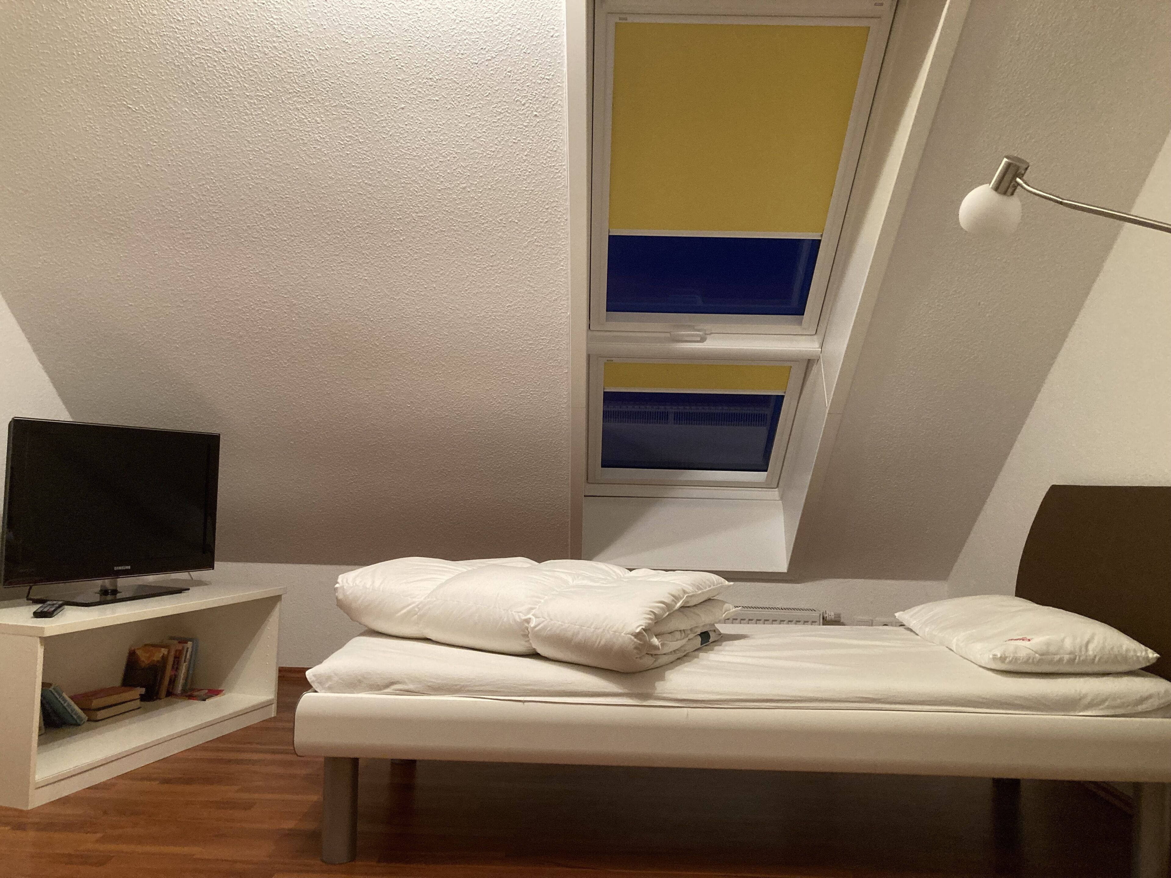 A bedroom with a bed, a TV on a stand, a window with yellow and blue blinds, and a lamp.