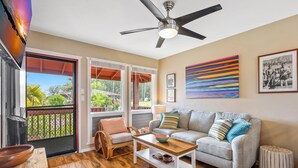 A living room with a grey sofa, wooden coffee table, wicker chair, and a ceiling fan. - Hale Moana E21, Coconut Grove Views, Walk to Kapaa Town and Beach (Kapaa)