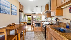 A modern kitchen with wooden cabinets, a granite countertop, and a built-in oven. There is a dining area with a wooden table and chairs. - Hale Moana E21, Coconut Grove Views, Walk to Kapaa Town and Beach (Kapaa)