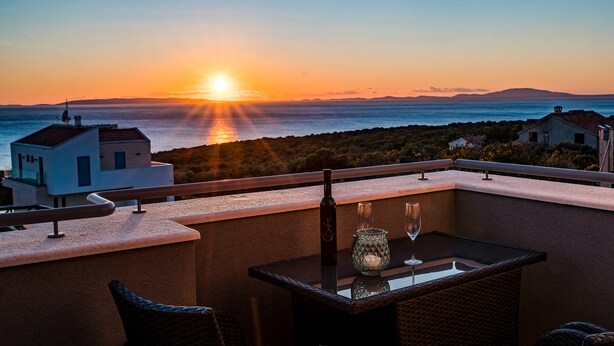 A balcony with a table, a bottle, and a glass, overlooking a sunset over the sea with buildings in the background. - Villa Dana with Private Pool (Novalja)