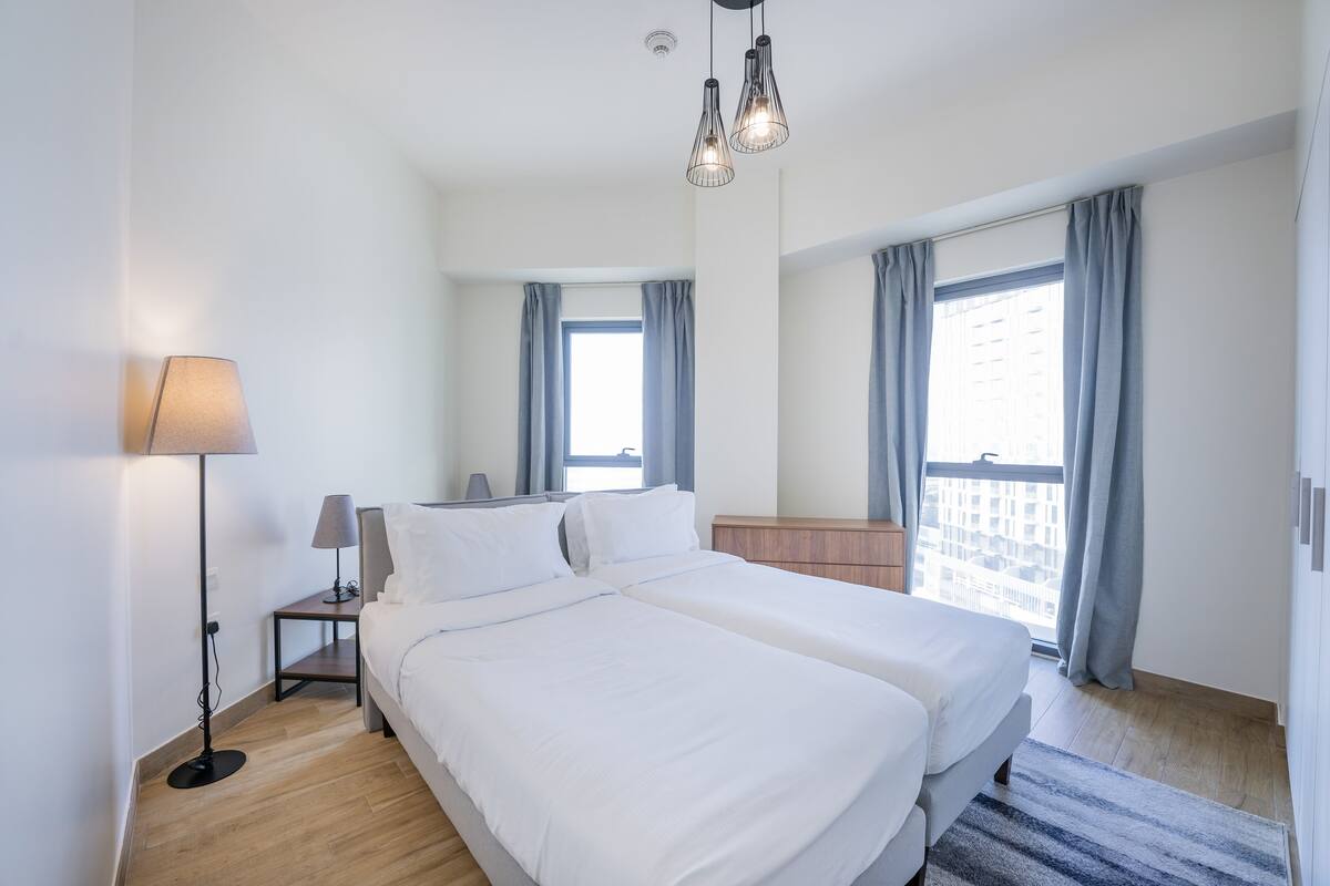 A modern hotel room with a large bed, a bedside table with a lamp, and a view of the city through large windows.