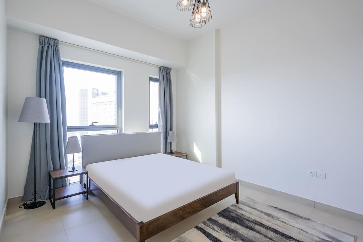 A bedroom with a large bed, two bedside tables with lamps, a window with curtains, and a chandelier.