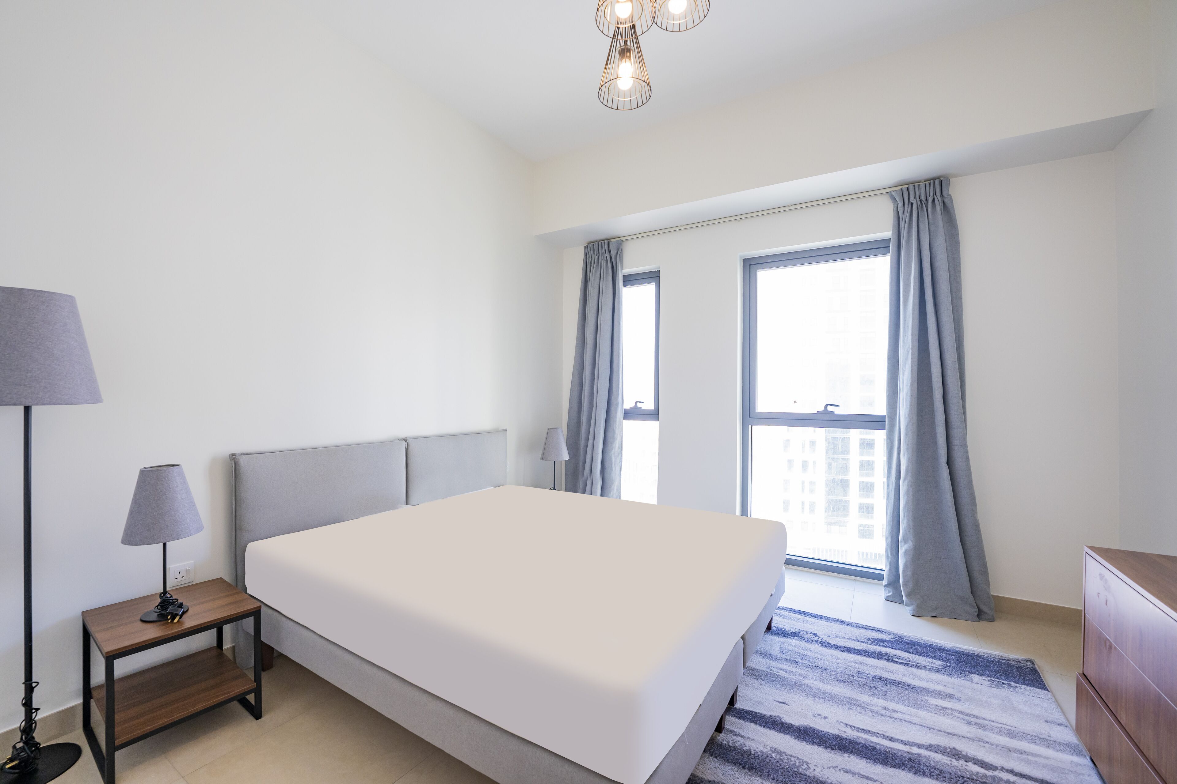 A modern hotel room with a large bed, a nightstand with a lamp, a dresser, and a view of the city through large windows.