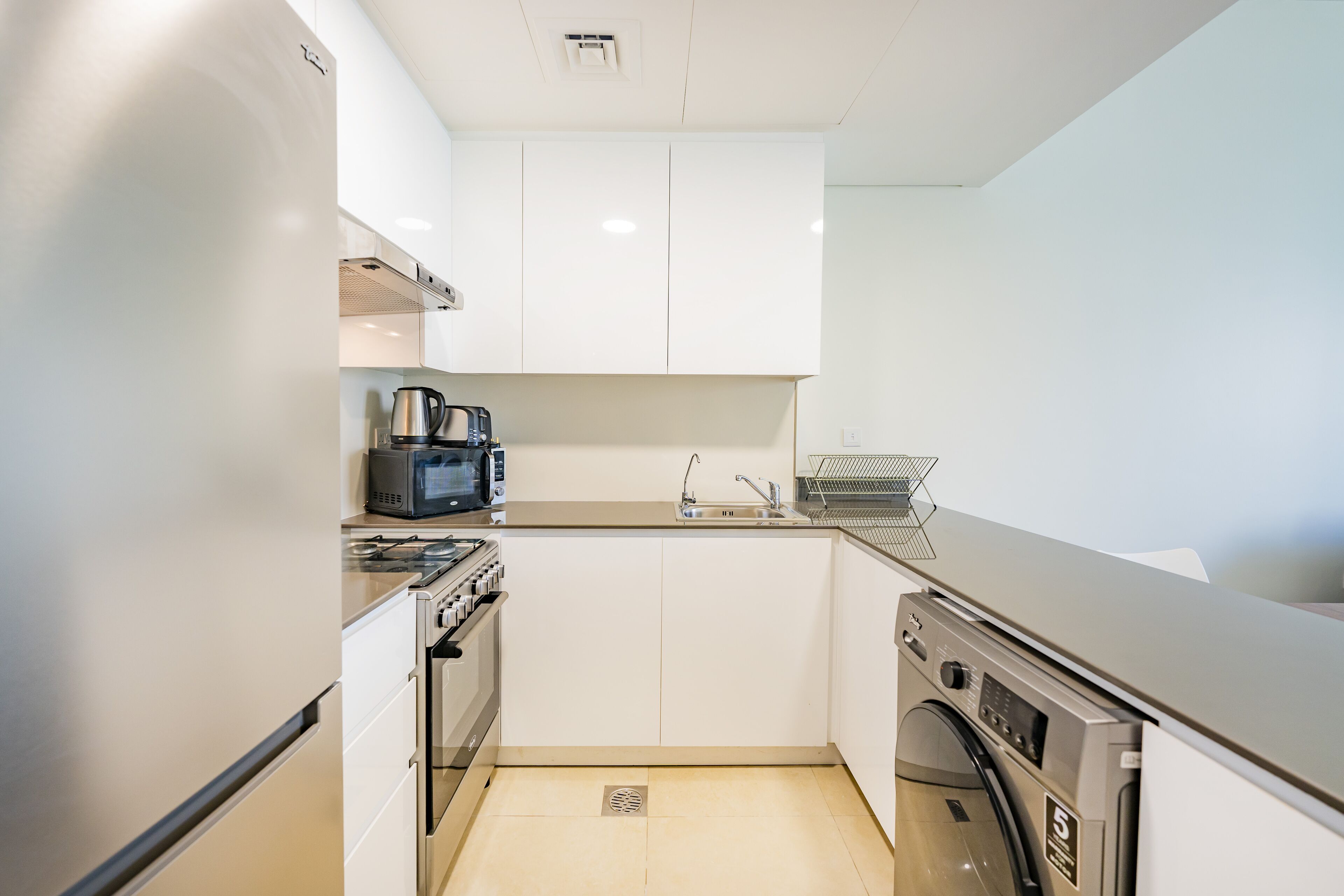 A compact kitchen with white cabinets, a microwave, a coffee maker, a sink, and a washing machine.