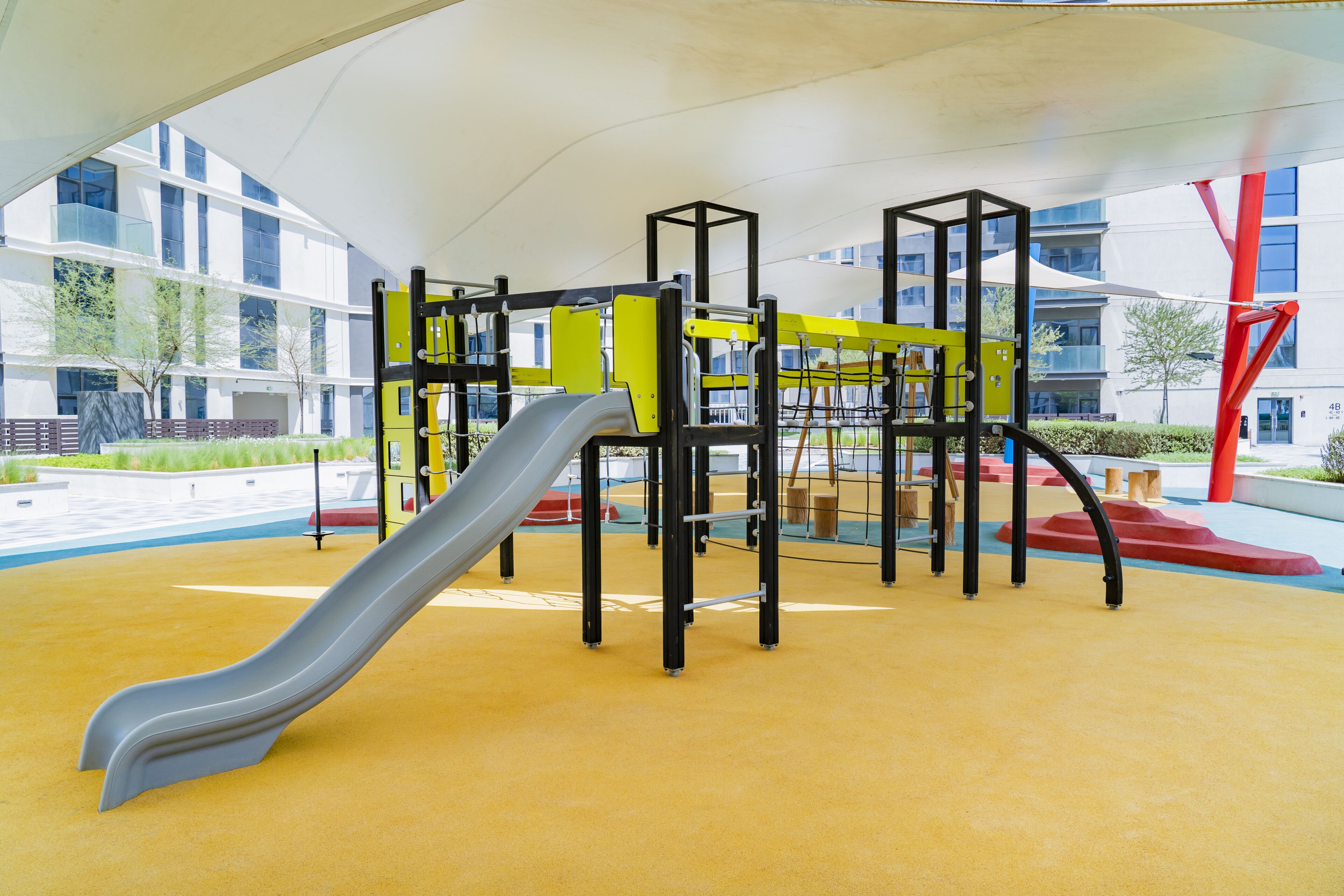 A playground with a slide and climbing structures, set against a modern building with large windows and a yellow play area.