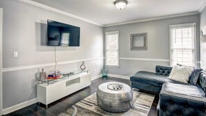 A modern living room with a flat-screen TV, a glass coffee table, a dark leather sofa, and a round ottoman. - Stone Mountain  Home  Amazing Yard & Pool (Stone Mountain)
