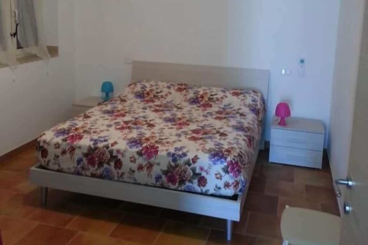 Une chambre à coucher avec un lit, une table de chevet, une chaise et une fenêtre avec des rideaux.