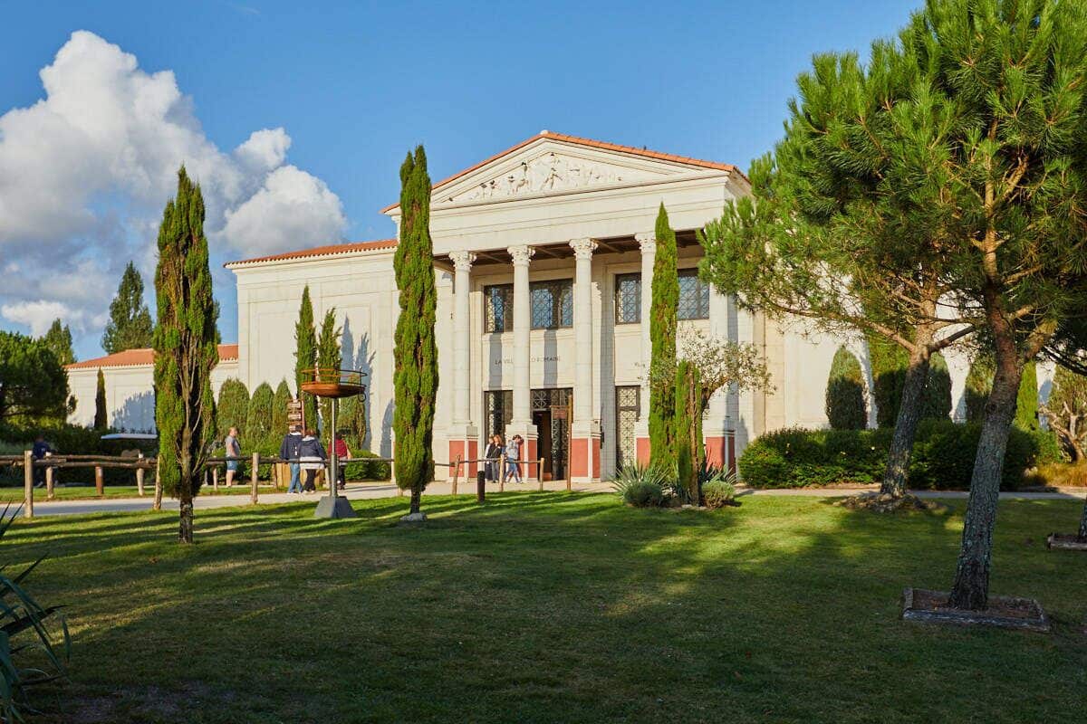 Un edificio clásico con columnas y un pórtico, rodeado de césped y árboles bien cuidados.