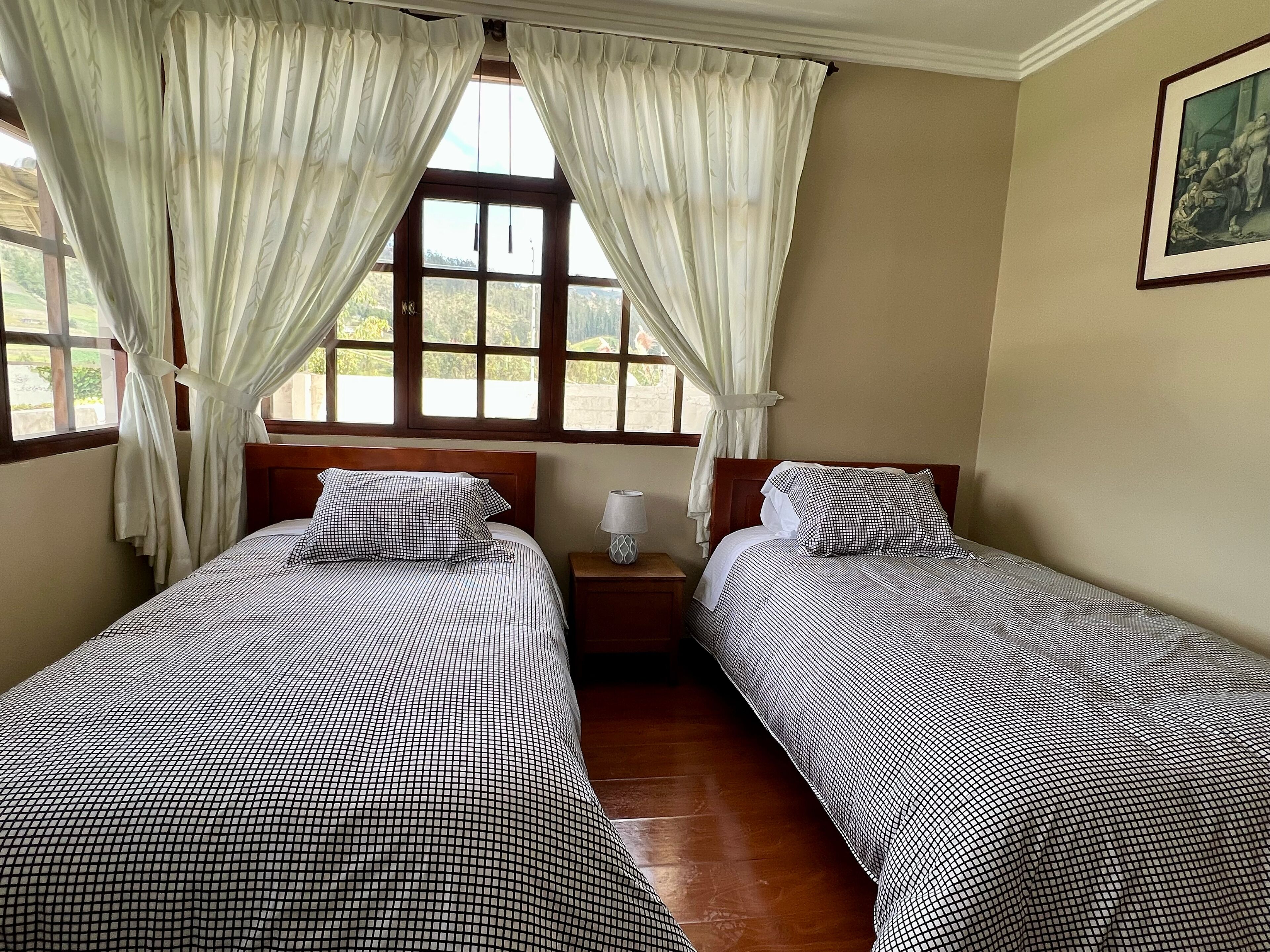 Una habitación con dos camas, una mesita de noche, una lámpara y un ventanal con cortinas.