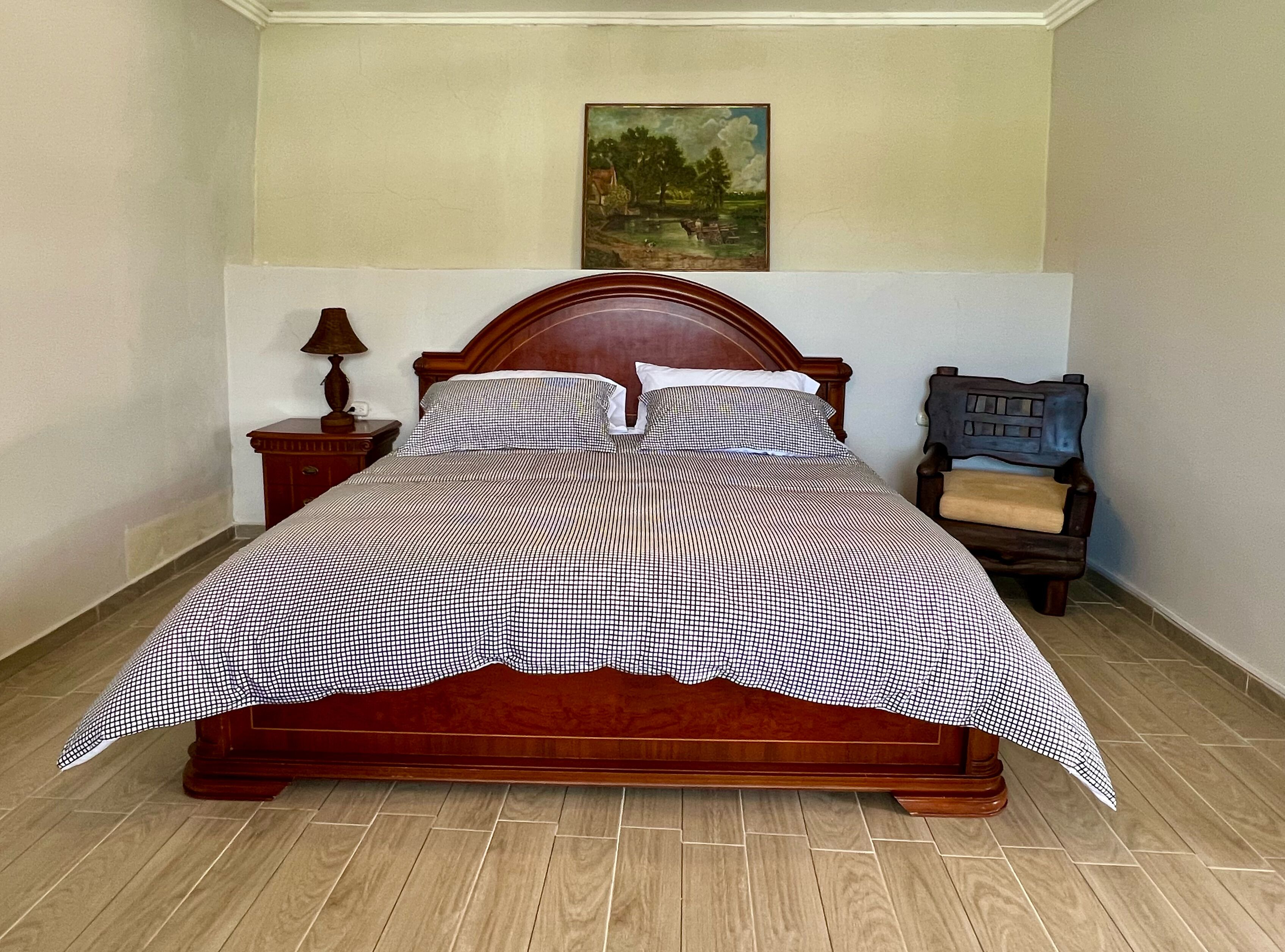 Una cama de madera con colcha a cuadros, una mesita de noche con lámpara, una silla y un cuadro en la pared.