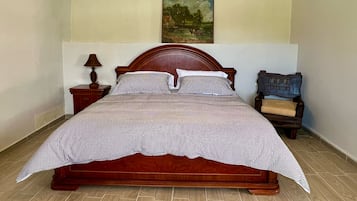 Una cama de madera con colcha a cuadros, una mesita de noche con lámpara, una silla y un cuadro en la pared.
