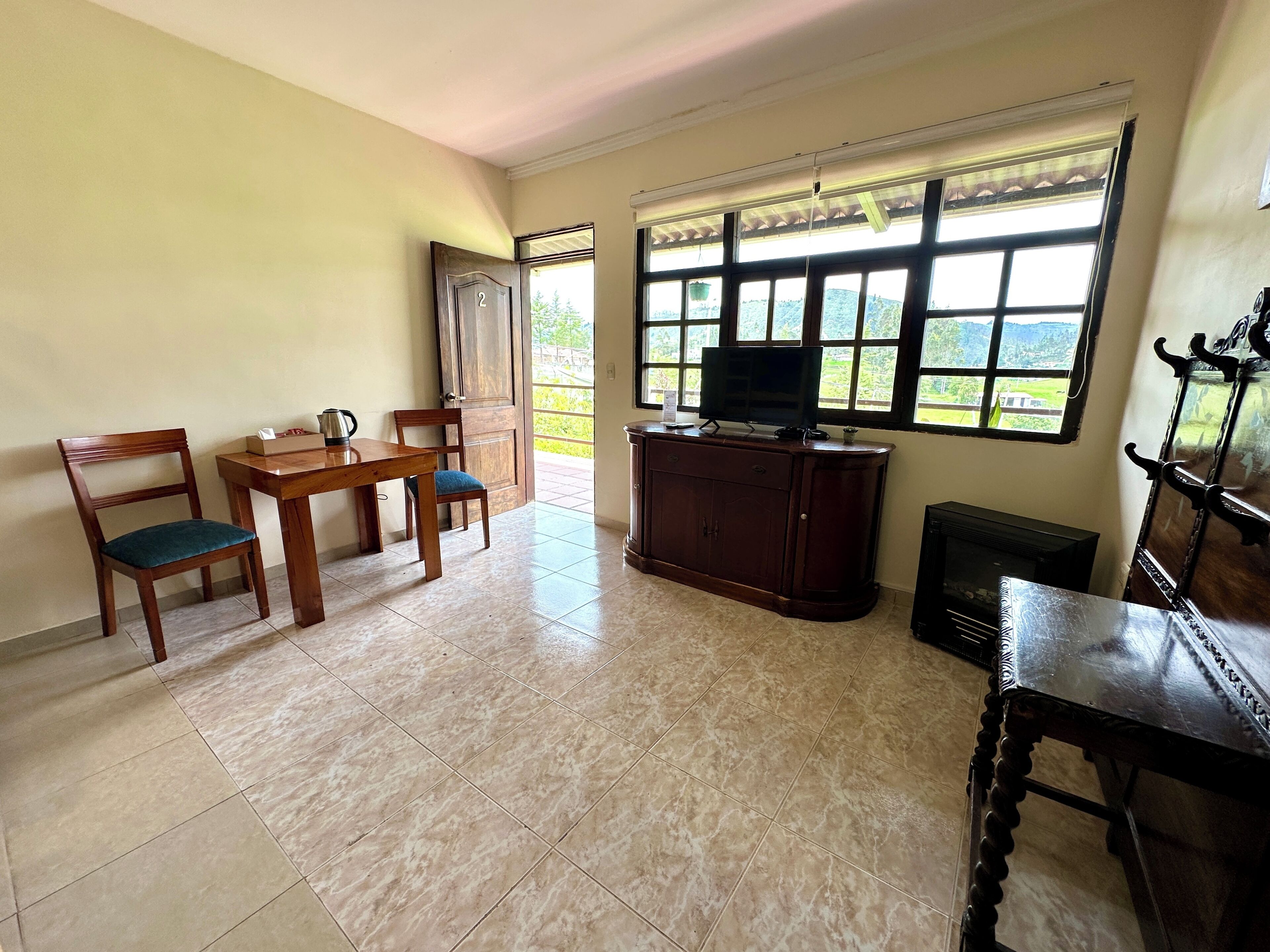 Una habitación con una mesa y sillas de madera, un televisor sobre un mueble, una chimenea y un ventanal con vistas a la vegetación.