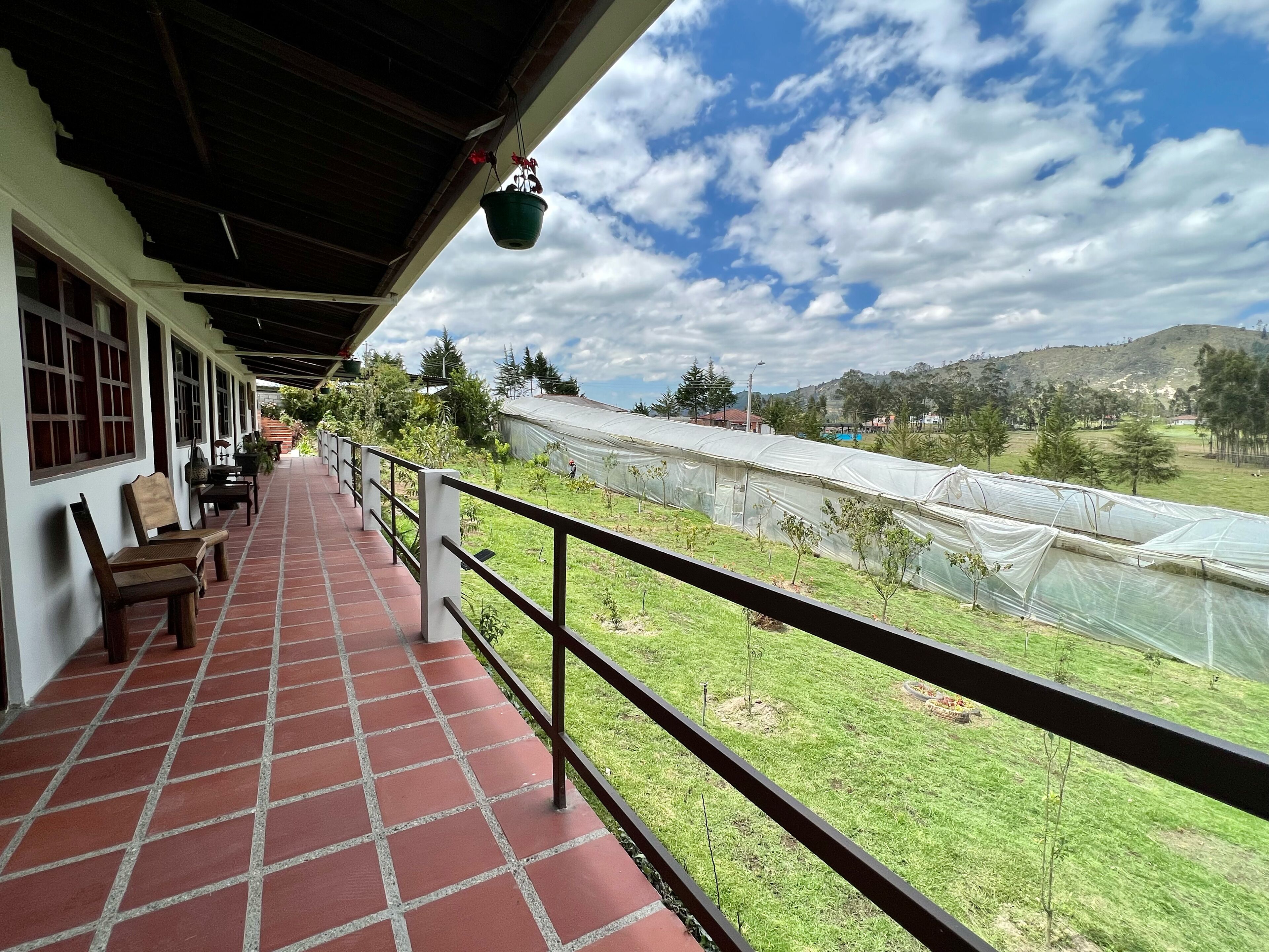 Un balcón cubierto con suelo de baldosas, barandilla y vistas a un invernadero y paisaje.
