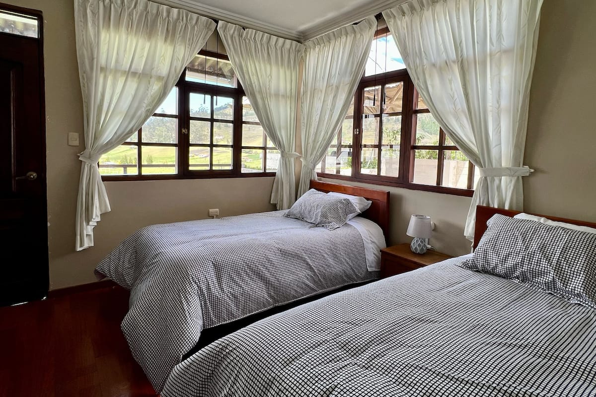 A room with two beds, white curtains, a wooden headboard, a bedside table with a lamp, and a window with a view of greenery.